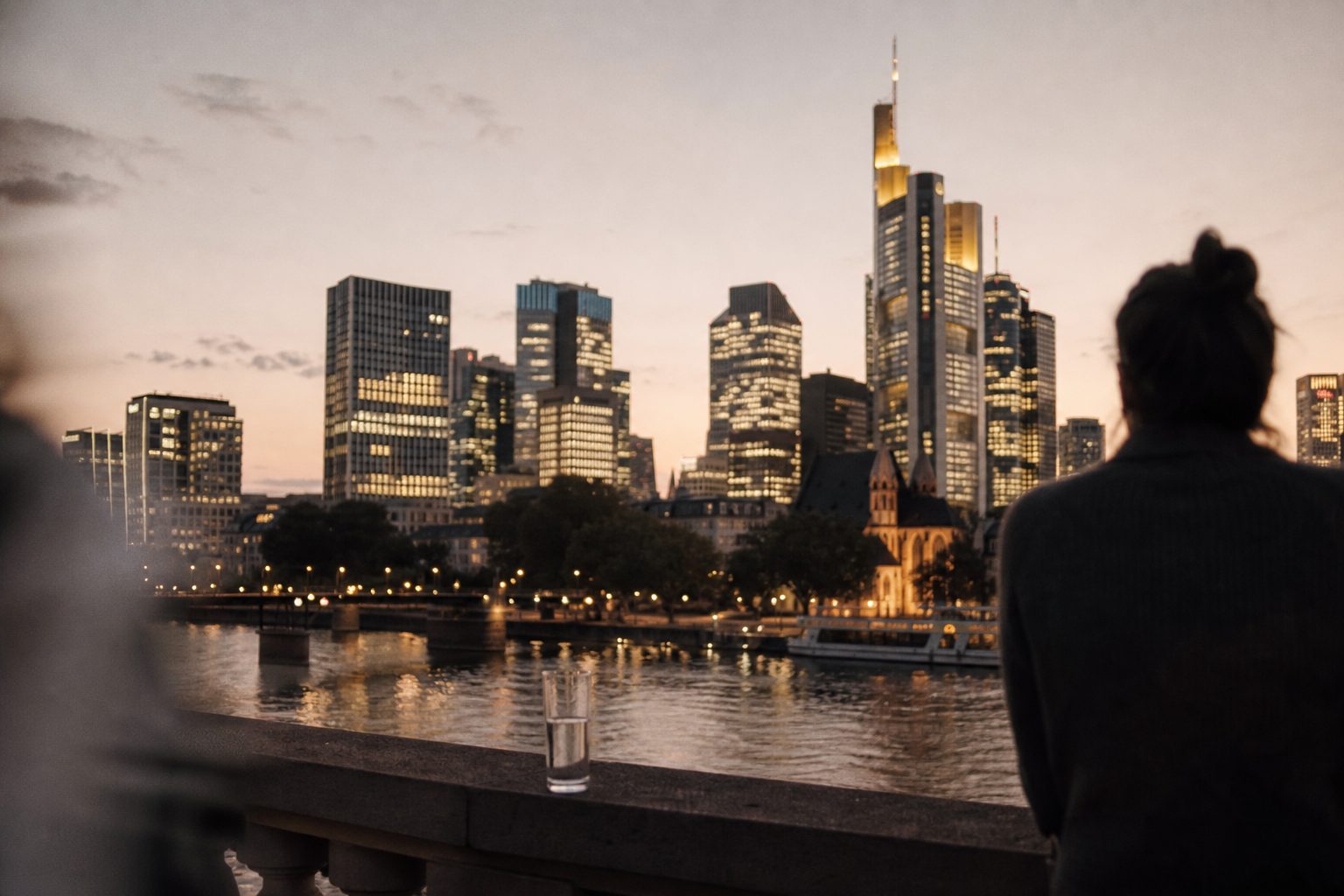Abendlicher Blick von einem Balkon auf die beleuchteten Bürotürme des Frankfurter Bankenviertels, im Vordergrund ein Glas Wasser auf der Brüstung.