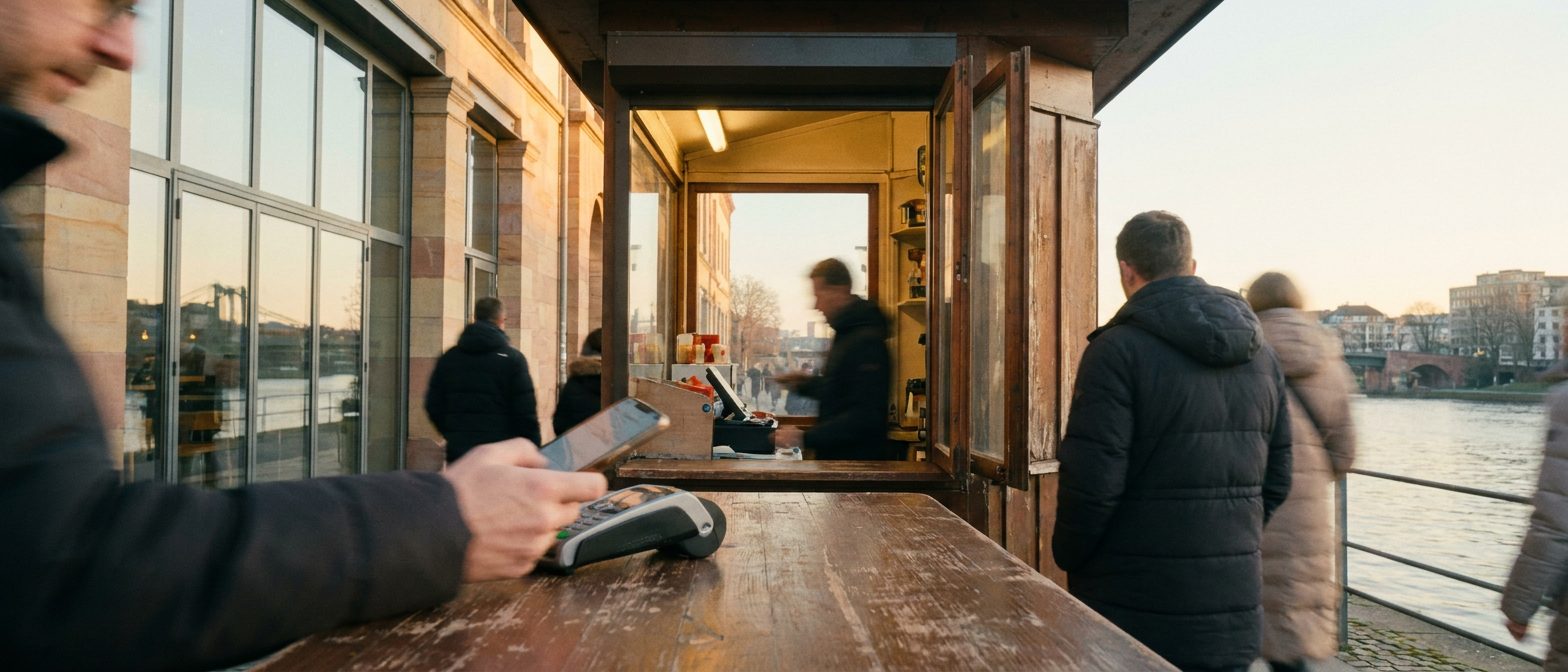 Eine Hand hält ein Smartphone an ein Kartenterminal auf einem Kiosktresen, im Hintergrund verschwimmen Regale.