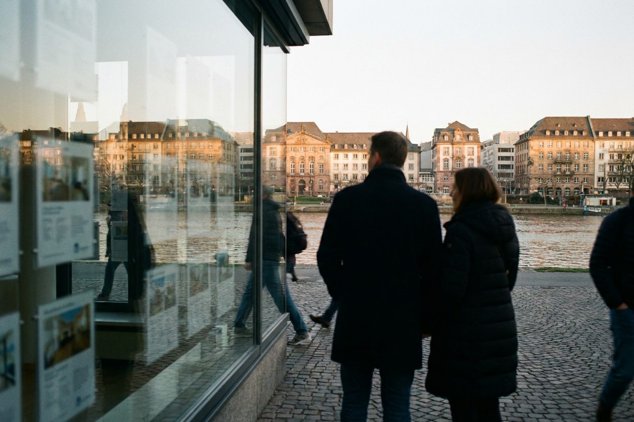 Ein Paar steht in der Dämmerung mit dem Rücken zur Kamera vor einem Schaufenster mit unscharfen Immobilienaushängen.