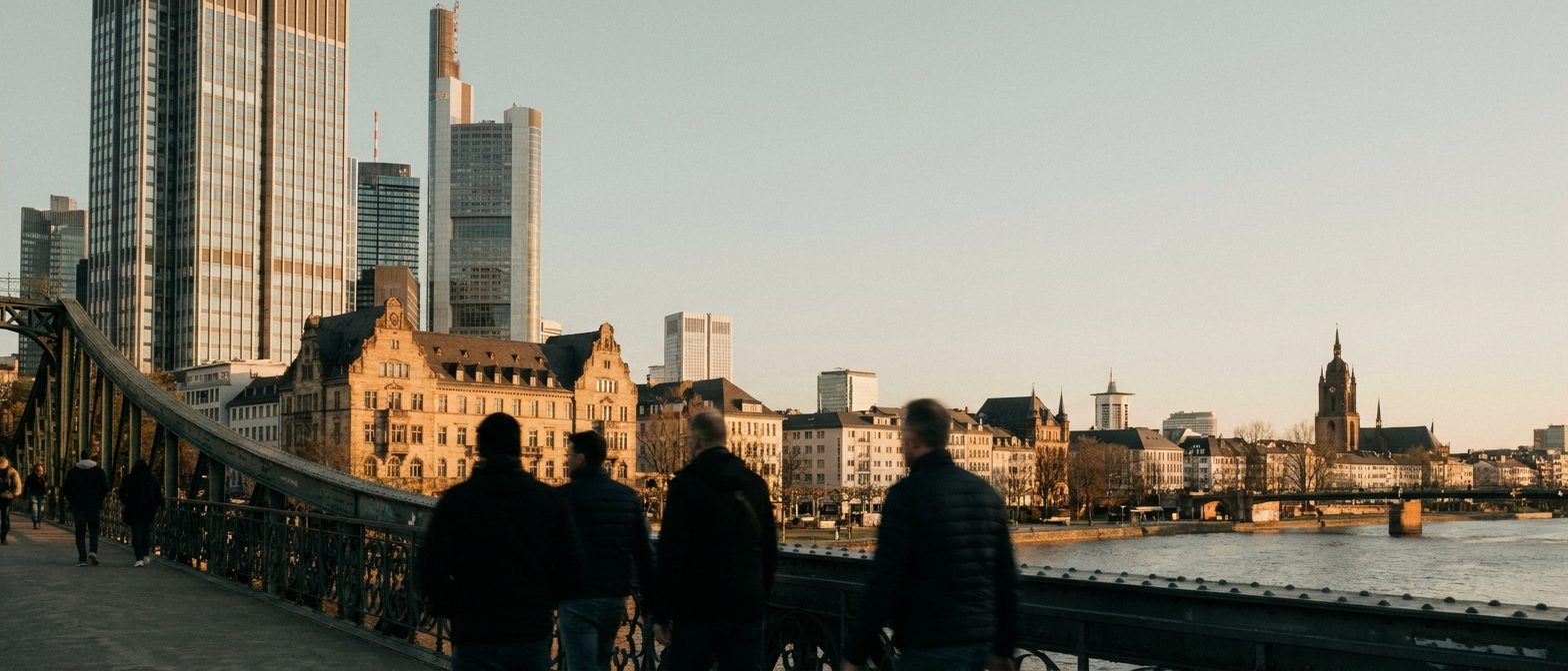 Blick vom Eisernen Steg auf die Frankfurter Skyline im warmen Nachmittagslicht, Passanten als Silhouetten auf der Brücke.