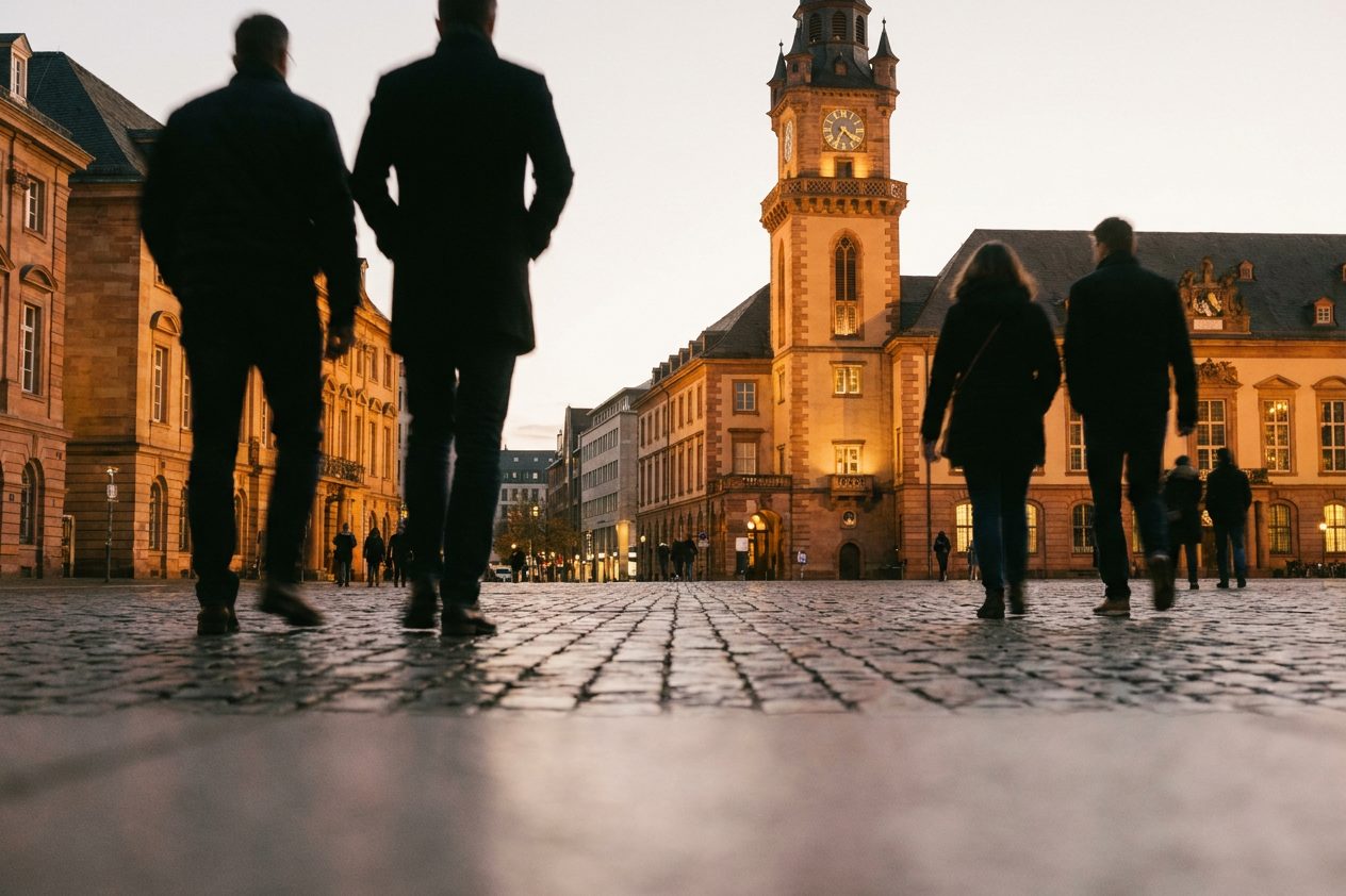 Passanten als Silhouetten auf dem Kaiserplatz in der Abenddämmerung vor warm beleuchteten Bankentürmen.